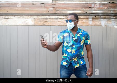 Ein ernsthafter junger Mann mit Gesichtsmaske und Sonnenbrille Und in ein Handy schauen Stockfoto