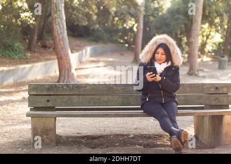 Eine junge hispanische Frau in einem Mantel sitzt auf einem Bank beim Telefonieren im Collserola Park Stockfoto