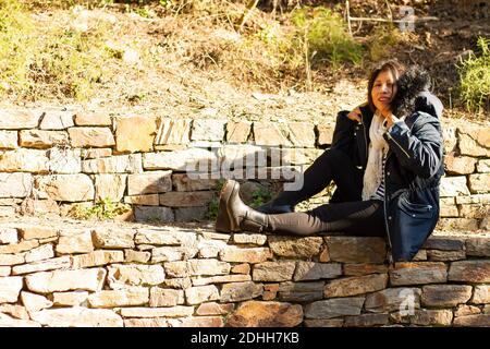 Eine junge hispanische Frau in einem Mantel sitzt auf einem Steinmauer im Collserola Park Stockfoto