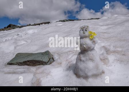 Kleine niedliche Schneemann mit einem Kranz von Blumen und ein Bouquet von polaren Mohnblumen auf dem Schnee hoch in der Berge vor dem Hintergrund von Berggipfeln Stockfoto