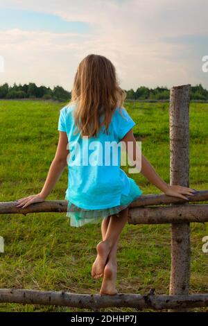 Ein kleines Mädchen ohne Schuhe sitzt auf einem Holzzaun Im Sommer auf einer Wiese Stockfoto