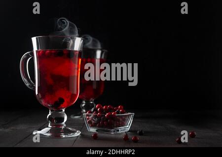 Weihnachtliche leuchtend rote Glühwein mit Beeren, Dampf, reife Preiselbeere, Heidelbeere in zwei Glas Weingläsern auf schwarzem Holztisch im dunklen Innenraum, Kopie sp Stockfoto