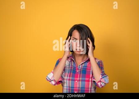 Eine Kopfschmerz habende Frau, die den Kopf hält, der versucht, Migräne zu behandeln, nach unten schaut, während sie gegen die gelbe Wand im Hintergrund mit Kopierraum steht Stockfoto