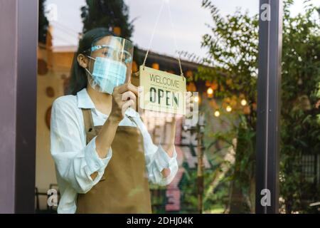 Asiatische Geschäftseigentümer Frau tragen Schutzmaske und Gesichtsschild hängen offen Zeichen in ihrem Restaurant / Café, wieder nach der Sperre für neue NOR öffnen Stockfoto