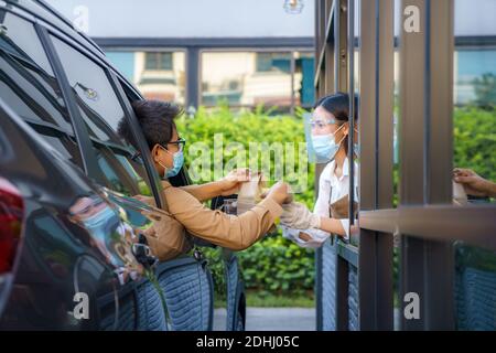 Asiatischer Mann in Schutzmaske unter Lebensmittelbeutel und Kaffee Mit Frau Kellnerin trägt Gesichtsmaske und Gesichtsschutz an Fahren Sie durch während der Coronavirus-Outbrea Stockfoto