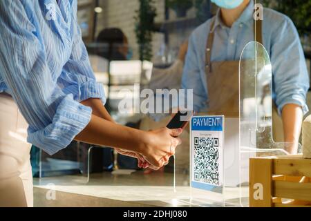 Asiatische Frau Kunden mit mobilen Scan QR oder NFC-Zahlung Auf dem Smartphone für ihr Kaffeegetränk im Kaffee bezahlen Shop und Plexi Bildschirm und Barista Wearin Stockfoto