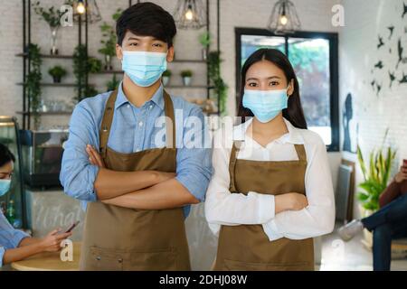 Asian Barista Paar in Schürze und Gesichtsmaske Blick auf Kamera bereit, Kaffee-Service an der modernen Kaffee geben Kaufen Stockfoto