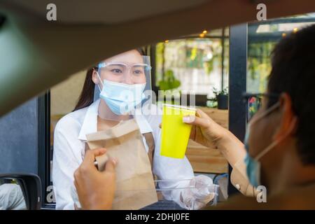 Asiatischer Mann in Schutzmaske unter Lebensmittelbeutel und Kaffee Mit Frau Kellnerin trägt Gesichtsmaske und Gesichtsschutz an Fahren Sie durch während der Coronavirus-Outbrea Stockfoto