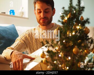 Kaukasischer junger Mann neben einem Weihnachtsbaum in einem Wollpullover, der ein Geschenk in den Händen hält. Er genießt die Ferien in einem gemütlichen Zuhause. Stockfoto