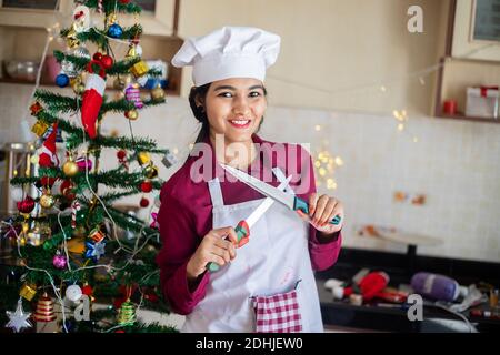 Portrait of Beautiful Young Indian Girl Chef trägt Schürze Holding Messer in der Hand posiert zu Hause Küche, Weihnachtsfeier und Vorbereitung concep Stockfoto