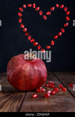 Ein frischer, reifer Granatapfel liegt auf einem braunen Brett auf schwarzem Hintergrund. Ein Herz aus Granatapfelkernen. Stockfoto
