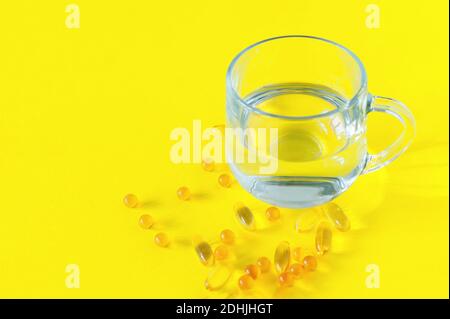 Fischölkapseln und Glas reines Wasser auf gelbem Hintergrund, Vitamin D, Omega-Ergänzung, selektiver Fokus. Stockfoto