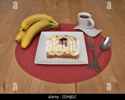 Eine Scheibe getoastetes Vollkornbrot, bedeckt mit einer Schicht Bananenbrötchen, garniert mit einem Löffel Himbeer. Platziert auf einer runden roten Matte b Stockfoto