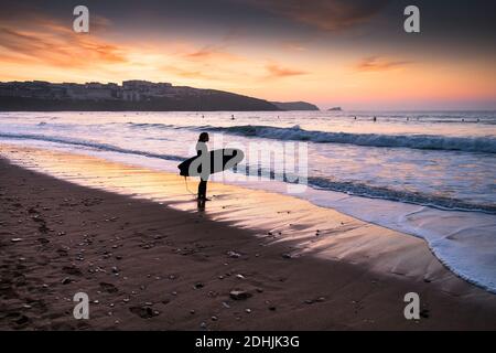 Ein Surfer, der am Strand von Fistral steht und vom Sonnenuntergang in Newquay in Cornwall umrahmt wird. Stockfoto
