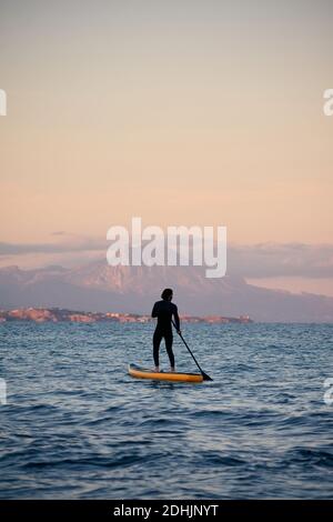 Männlicher Surfer im Neoprenanzug Rudern auf dem Paddelbrett auf dem Meer Wasser auf dem Hintergrund der Berge bei Sonnenuntergang Stockfoto