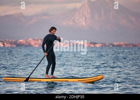 Seitenansicht des männlichen Surfers im Neoprenanzug, der auf dem Paddel rudert Bord auf dem Meerwasser auf dem Hintergrund der Berge bei Sonnenuntergang Stockfoto