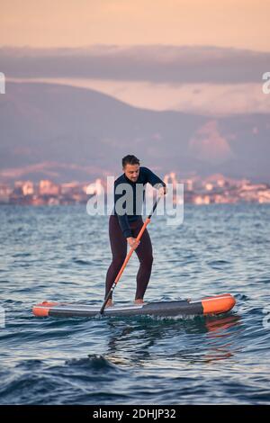 Männlicher Surfer im Neoprenanzug Rudern auf dem Paddelbrett auf dem Meer Wasser auf dem Hintergrund der Berge bei Sonnenuntergang Stockfoto