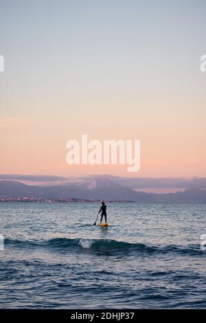 Männlicher Surfer im Neoprenanzug Rudern auf dem Paddelbrett auf dem Meer Wasser auf dem Hintergrund der Berge bei Sonnenuntergang Stockfoto