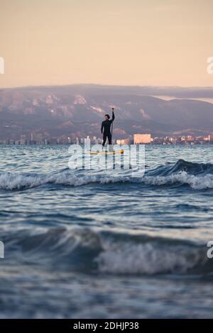 Männlicher Surfer im Neoprenanzug Rudern auf dem Paddelbrett auf dem Meer Wasser auf dem Hintergrund der Berge bei Sonnenuntergang Stockfoto