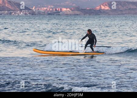 Männlicher Surfer im Neoprenanzug Rudern auf dem Paddelbrett auf dem Meer Wasser auf dem Hintergrund der Berge bei Sonnenuntergang Stockfoto