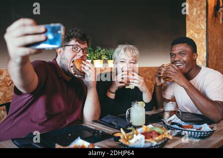Fröhliche multiethnische Freunde essen Burger in Fast-Food-Café und Selfie beim Sammeln und Spaß haben Stockfoto