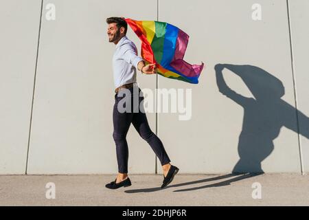 Seitenansicht der fröhlichen homosexuellen männlichen im Moment des Sprüngens Mit Regenbogen-LGBT-Flagge auf der Stadtstraße an sonnigen Tagen Stockfoto