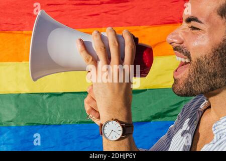 Beschnitten Seitenansicht aufgeregt homosexuellen männlichen stehend in der Nähe von Gebäude Mit LGBT-Regenbogenfahne und Schreien in Lautsprechern Stockfoto