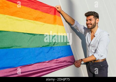 Seitenansicht des fröhlichen homosexuellen Mannes mit LGBT-Flagge in Regenbogen Stehen auf Betonmauer auf der Stadtstraße an sonnigen Tag Stockfoto