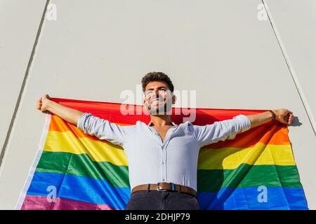 Von unten fröhlich homosexuellen Mann mit Regenbogen LGBT Flagge stehen An der Betonmauer auf der Stadtstraße am sonnigen Tag Stockfoto