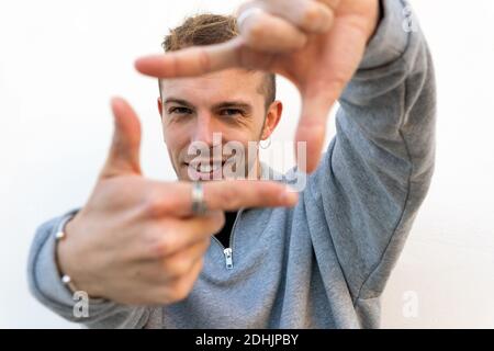 Lächelnd junge Hipster Kerl in lässigen Outfit machen Fotorahmen Mit den Fingern und Blick auf die Kamera vor weißem Hintergrund Stockfoto