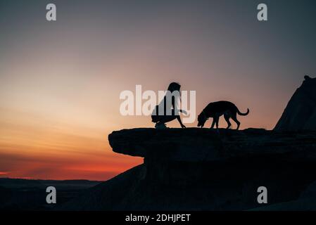 Seitenansicht der Silhouette von nicht erkennbaren weiblichen sitzen auf Hügel Mit Hund auf dem Hintergrund der Sonnenuntergang in Bardenas Reales Stockfoto