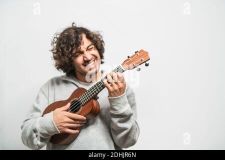 Positive junge Curly haired ethnischen Musiker in Casual tragen Durchführung Musik auf Ukulele-Gitarre gegen weiße Wand Stockfoto