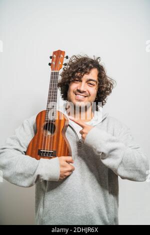 Positive junge Curly haired ethnischen Musiker in Casual tragen Durchführung Musik auf Ukulele Gitarre gegen weiße Wand Blick auf die Kamera Stockfoto