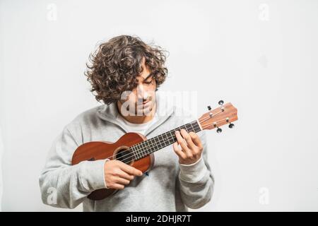 Positive junge Curly haired ethnischen Musiker in Casual tragen Durchführung Musik auf Ukulele-Gitarre gegen weiße Wand Stockfoto