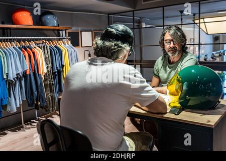 Seitenansicht von zwei Personen, die an einem Holztisch sitzen Beim Gespräch neben einem Motorradhelm in einer Kleidung Speichern Stockfoto
