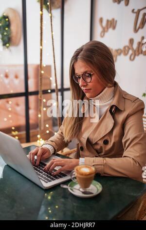Junge Bloggerin in legerer Kleidung und Brille sitzen an Tisch mit Tasse Kaffee und Tastatur auf Laptop-Tastatur Beim Vorbereiten von Inhalten für Socia Stockfoto