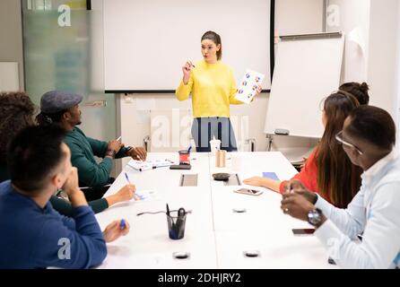 Fokussierte, multirassische Kollegen sitzen um den Tisch herum und hören einer Sprecherin zu Während der Präsentation und Diskussion der Geschäftsstrategie in modernen Workp Stockfoto