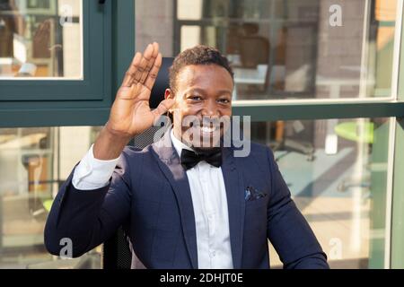 Fröhliche Erwachsene afroamerikanische männliche Unternehmer in eleganten formellen Anzug Mit Fliege Blick auf Kamera und winkende Hand während Sitzen gegen Glas wa Stockfoto