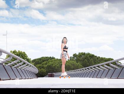 Voller Körper fit junge Frau in trendigen activewear Skating Auf Longboard entlang eingezäunten Brücke während der Ausübung aktiven Lebensstil in Sommerzeit Stockfoto