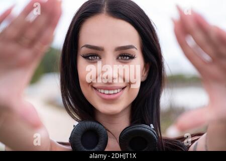 Attraktive junge Brünette mit stilvollen Make-up und Kopfhörer am Hals Bilder mit den Händen erstellen und mit einem Lächeln auf die Kamera schauen Stockfoto