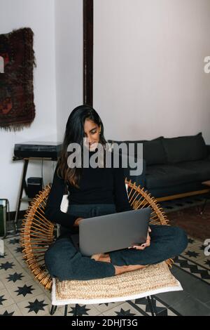Indische Freiberuflerin sitzt mit gekreuzten Beinen in Korbstuhl Und beim Online-Arbeiten von zu Hause aus auf Netbook tippen Stockfoto