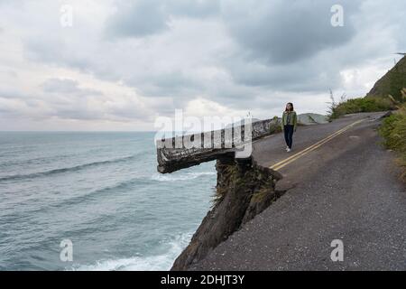 Nicht erkennbare entfernt asiatische ethnische weibliche Reisende am Rande der Zerstörte asphaltierte Straße entlang rauer felsiger Küste in der Nähe von winken Meer in rüber Stockfoto