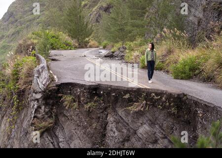 Nicht erkennbare entfernt asiatische ethnische weibliche Reisende am Rande der Zerstörte asphaltierte Straße entlang rauer felsiger Küste in der Nähe von winken Meer in rüber Stockfoto