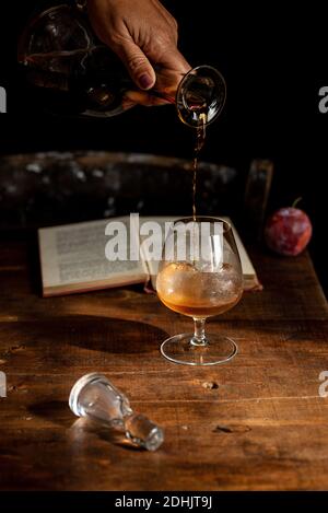 Crop anonyme Person Gießen Cognac aus Dekanter in Glas platziert Auf dem Holztisch neben dem offenen Buch Stockfoto