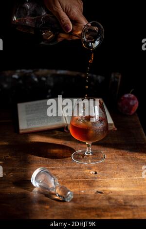 Crop anonyme Person Gießen Cognac aus Dekanter in Glas platziert Auf dem Holztisch neben dem offenen Buch Stockfoto