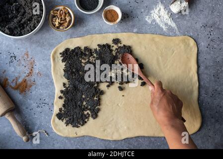 Oben der Ernte anonyme Person mit dem hölzernen Löffel, der sich ausbreitet mohnsamen auf gerolltem Teig während der Zubereitung von süßem Teig Tisch mit Zutaten Stockfoto