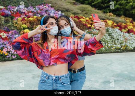 Fröhliche Freundinnen in trendigen Outfits und Schutzmasken hören Musik mit Kopfhörern und Selfie auf dem Smartphone beim Genießen Sommer d Stockfoto