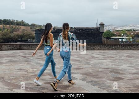 Seitenansicht von jungen schlanken Freundinnen in trendigen Outfits und Schutzmasken, die während des Coronavirus auf dem gepflasterten Stadtplatz zusammen laufen Ausbruch Stockfoto