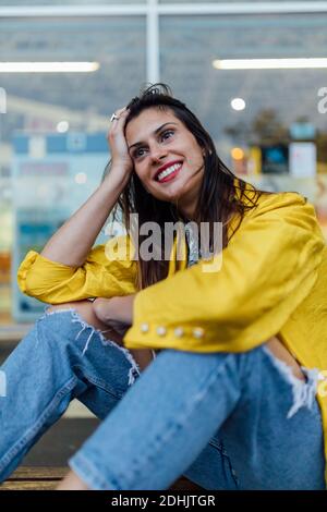 Junge langhaarige Frau in trendigen gelben Lederjacke und Zerrissene Jeans schaut weg, während sie auf der Bank gegen Glas sitzt Fenster eines modernen Gebäudes Stockfoto