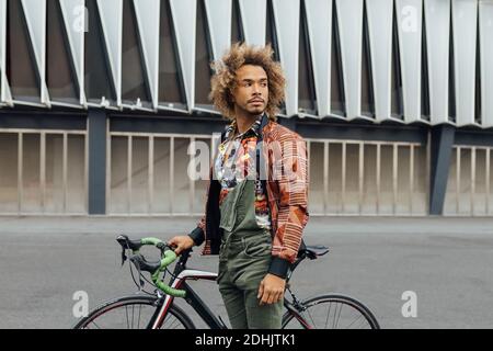 Junge Hipster Curly haired African American männlich in trendigen tragen Stehen mit Fahrrad auf dem Stadtplatz gegen moderne Gebäude suchen Weg Stockfoto